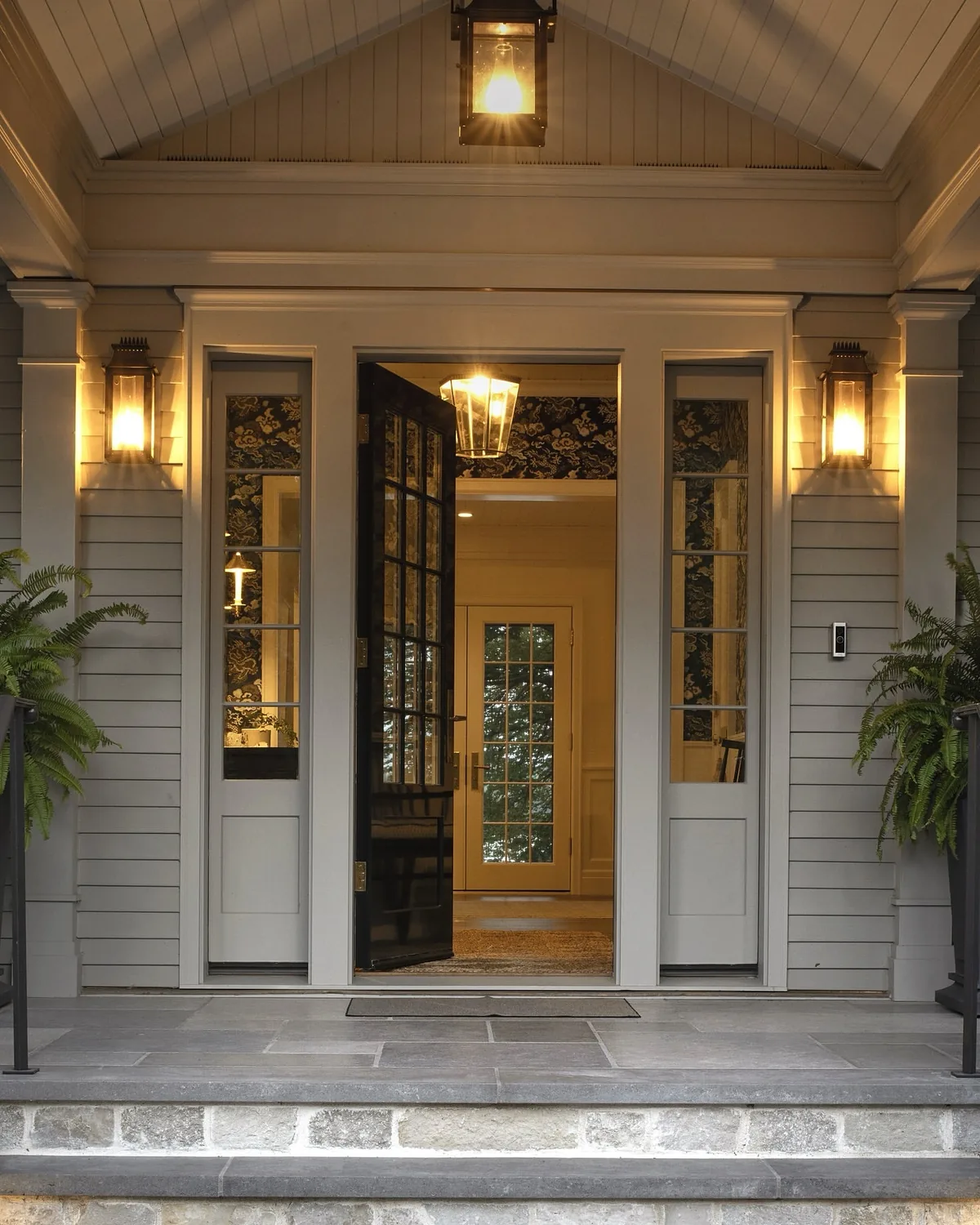 Gray clapboard portico with a glass front door glowing behind