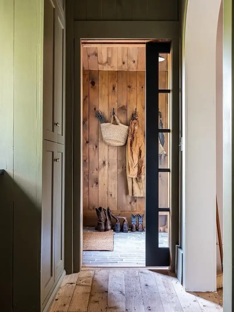 Rustic mudroom entry with knotty pine walls, wrought-iron hooks, straw tote, and a row of cowboy boots
