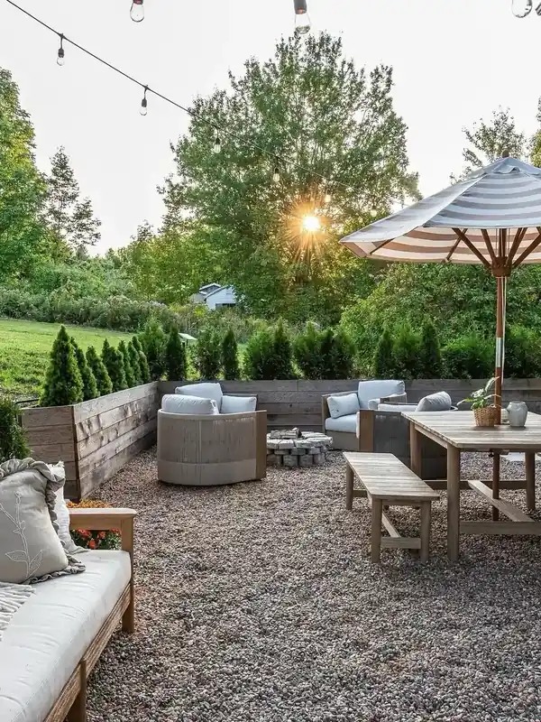 Gravel patio outdoor living room with wood furniture, a stone fire pit, and string lights at sunset