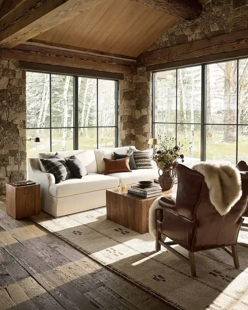 Rustic living room with natural stone walls, exposed wood beams, cream linen sofa, and black-framed windows overlooking a snowy forest