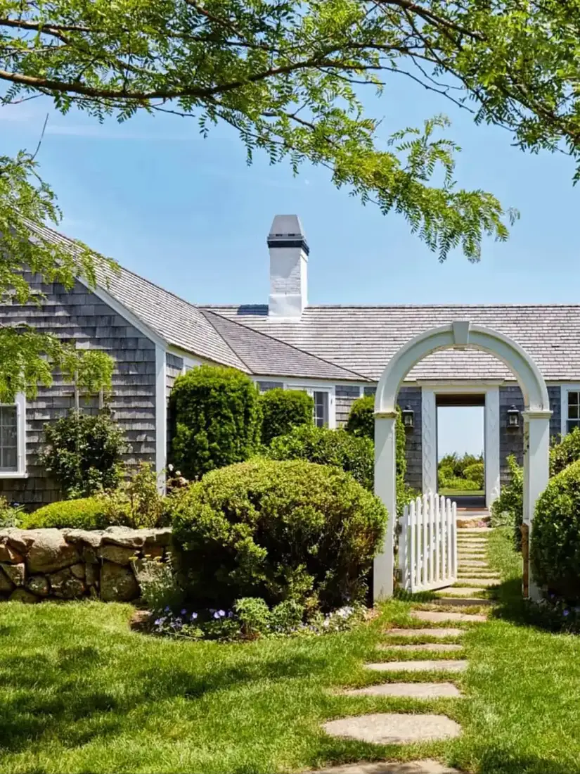 Grey weathered shingle cottage with a white arbor gate at the garden entrance