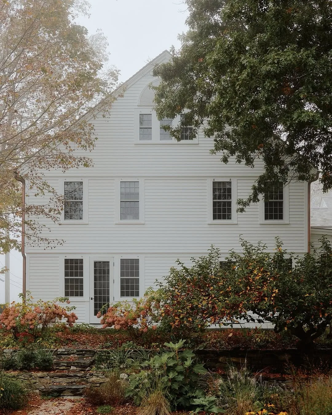 White clapboard gable with a shingle roof and garden beds