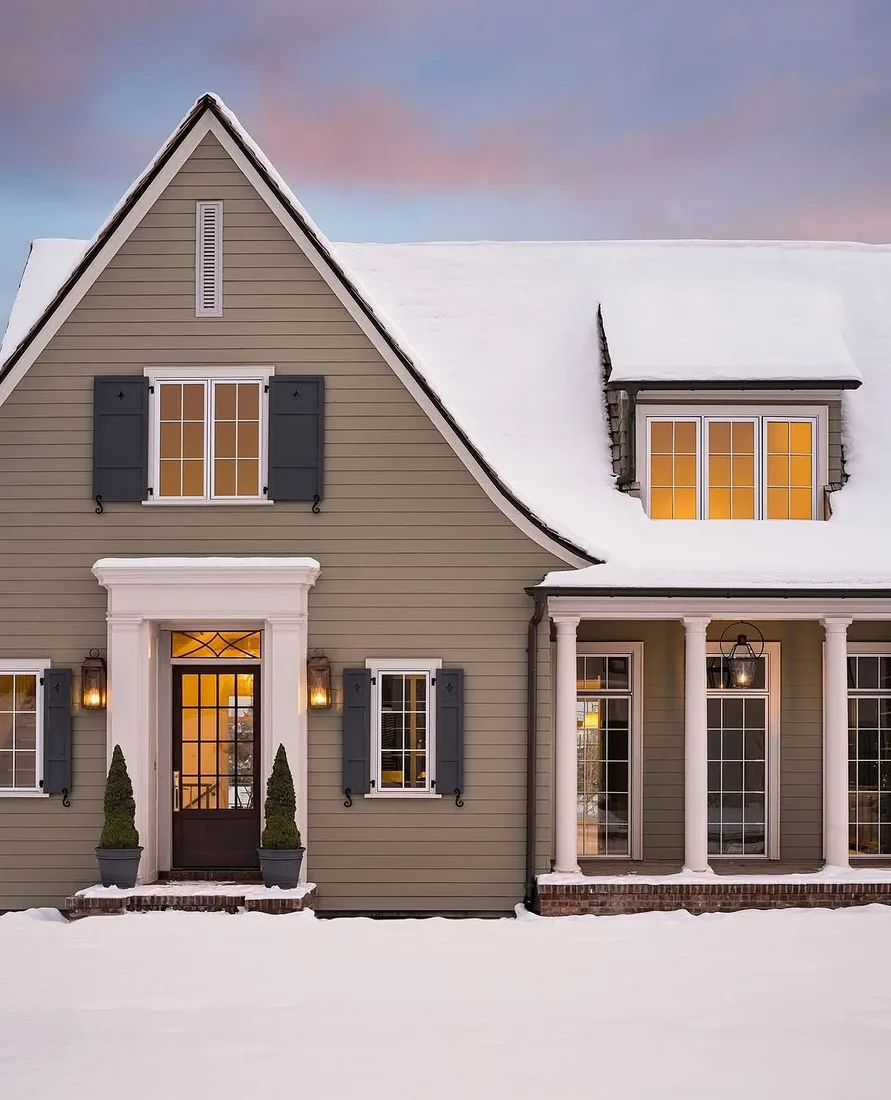 Grey shingle house exterior in snow at evening with warm window light
