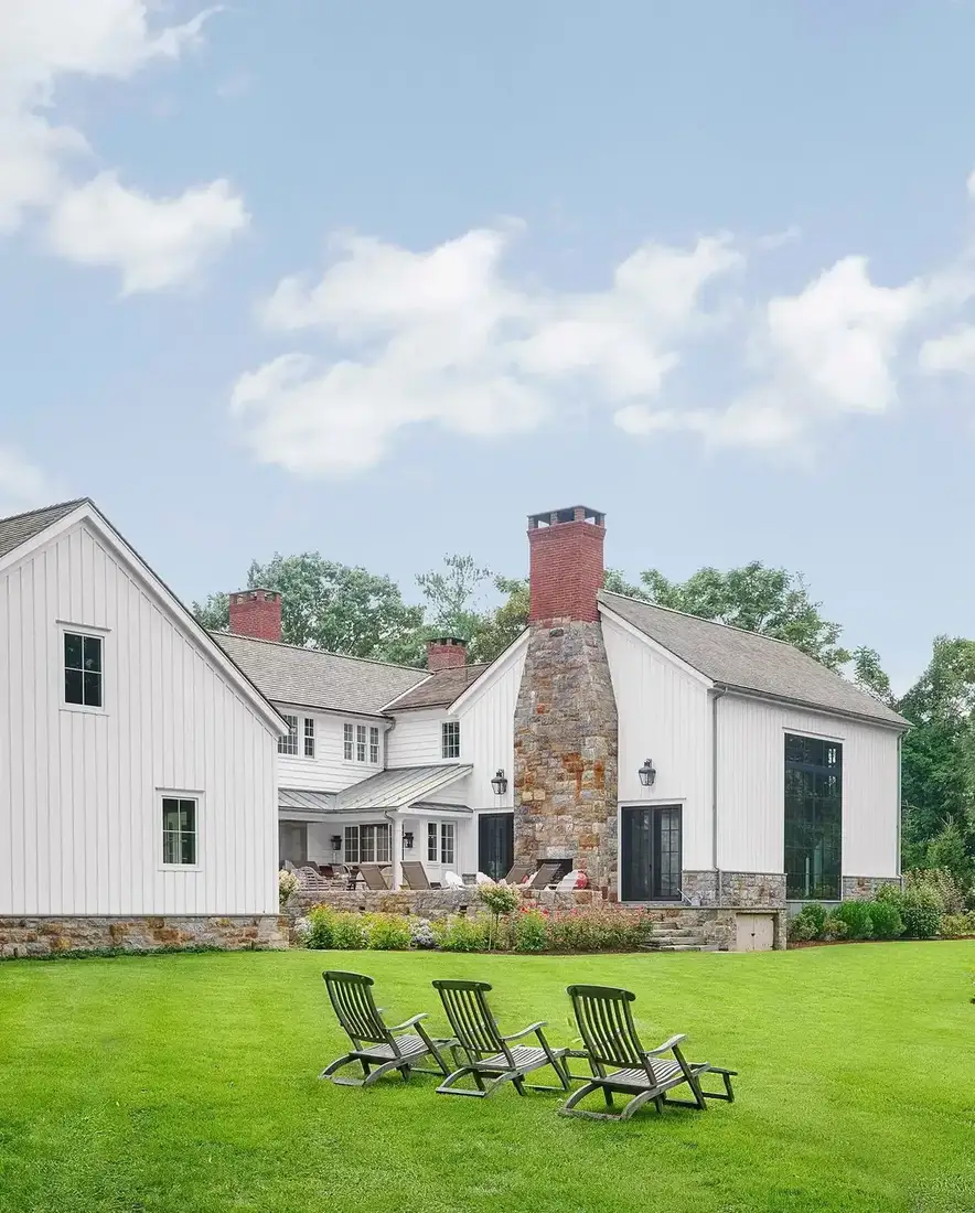 White board and batten farmhouse with stone chimney and wide sloping lawn
