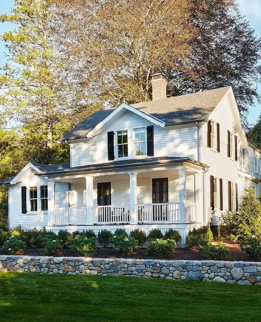 Cream farmhouse with wraparound porch and low stone wall along the front