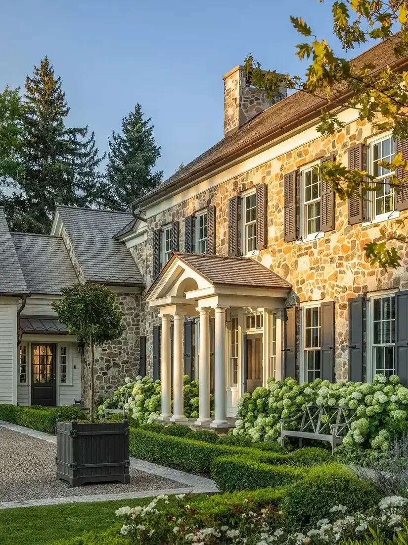 Fieldstone farmhouse with black shutters and clipped boxwood along the front path