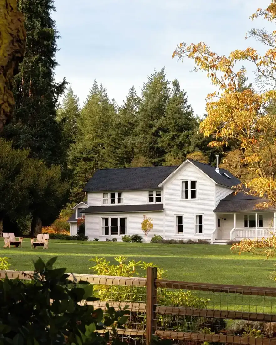 White farmhouse with a black metal roof set behind a post and rail fence in an open meadow